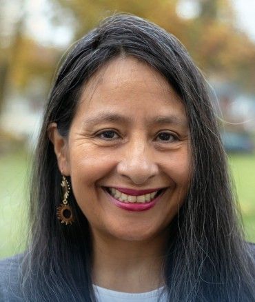Woman smiling outdoors with long dark hair, wearing earrings and a blue blazer.