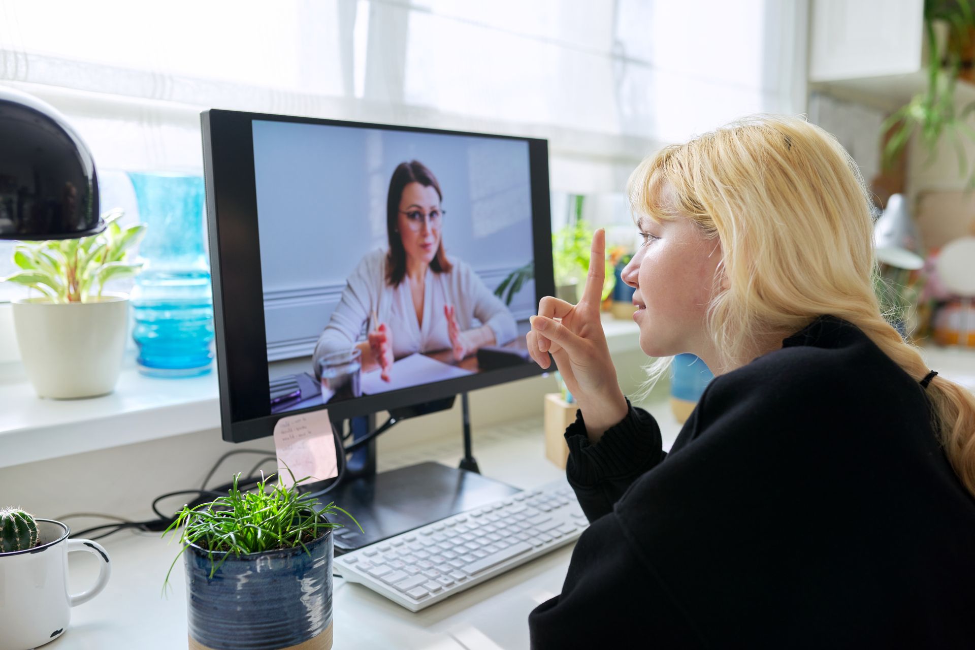 Woman on a video call, pointing. Another woman on the screen, looking at her, papers on desk. Desktop computer.