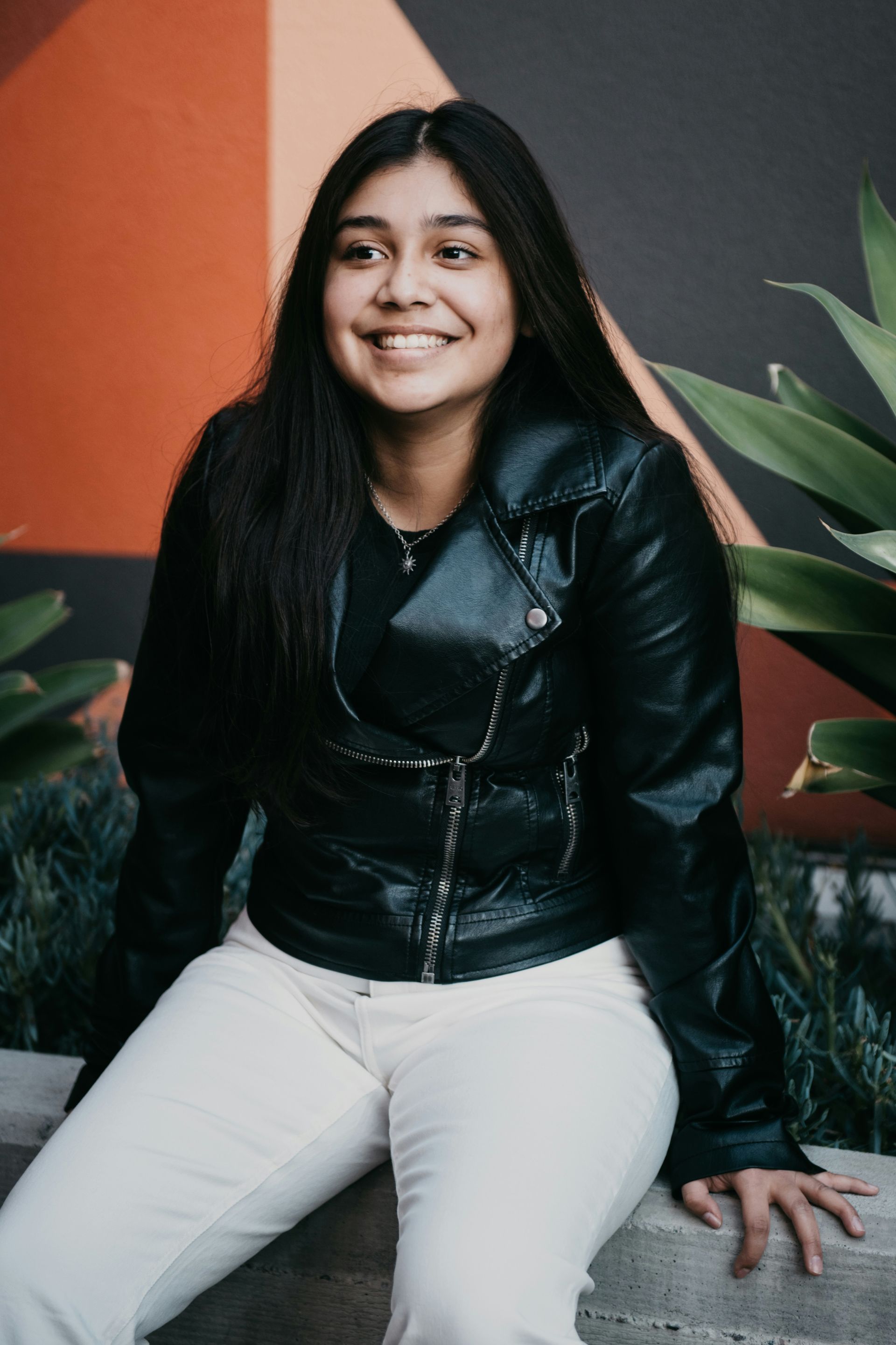 Woman in black jacket and white pants, smiling, seated outdoors.