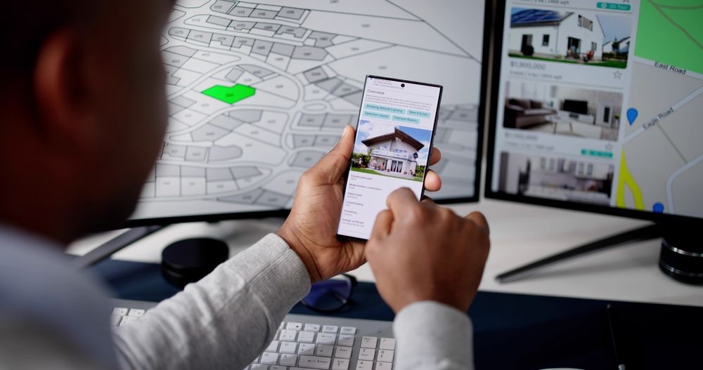 A man is holding a cell phone in front of a computer screen.