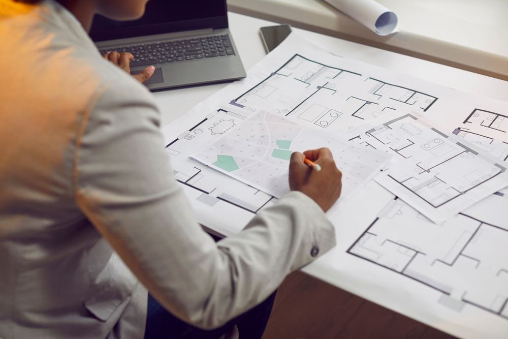 A woman is sitting at a desk with a laptop and a blueprint of a house.