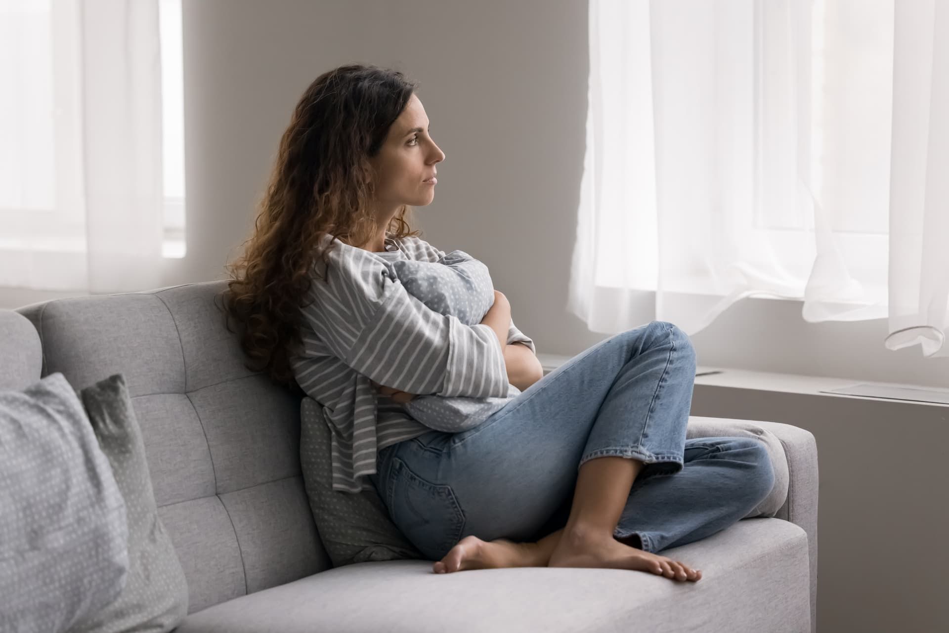 Woman sits on a couch, looking out a window. She is holding a pillow and appears thoughtful.