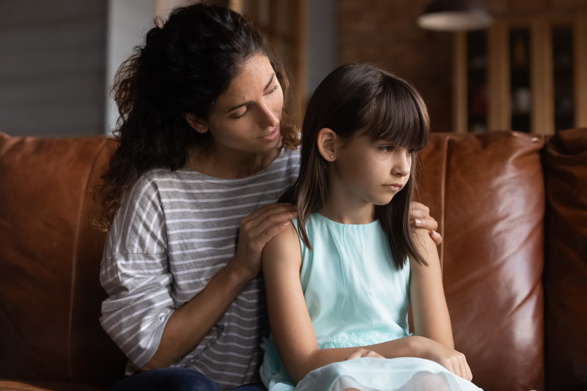 Woman consoles a girl on a brown sofa; woman's hand on girl's shoulder. Girl looks sad.