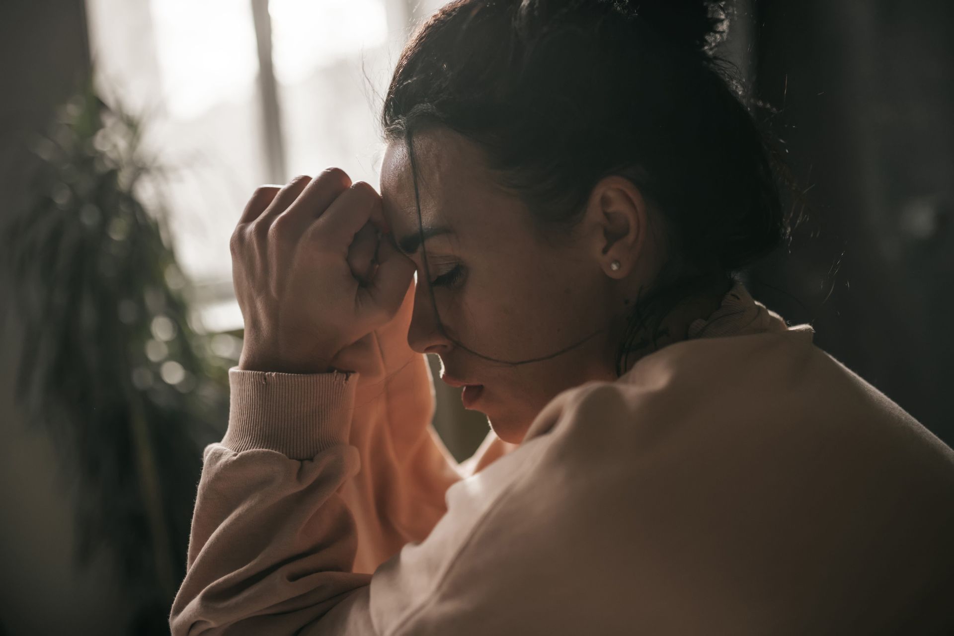 Woman in a peach sweater rests her head on her hand by a sunlit window, looking pensive.