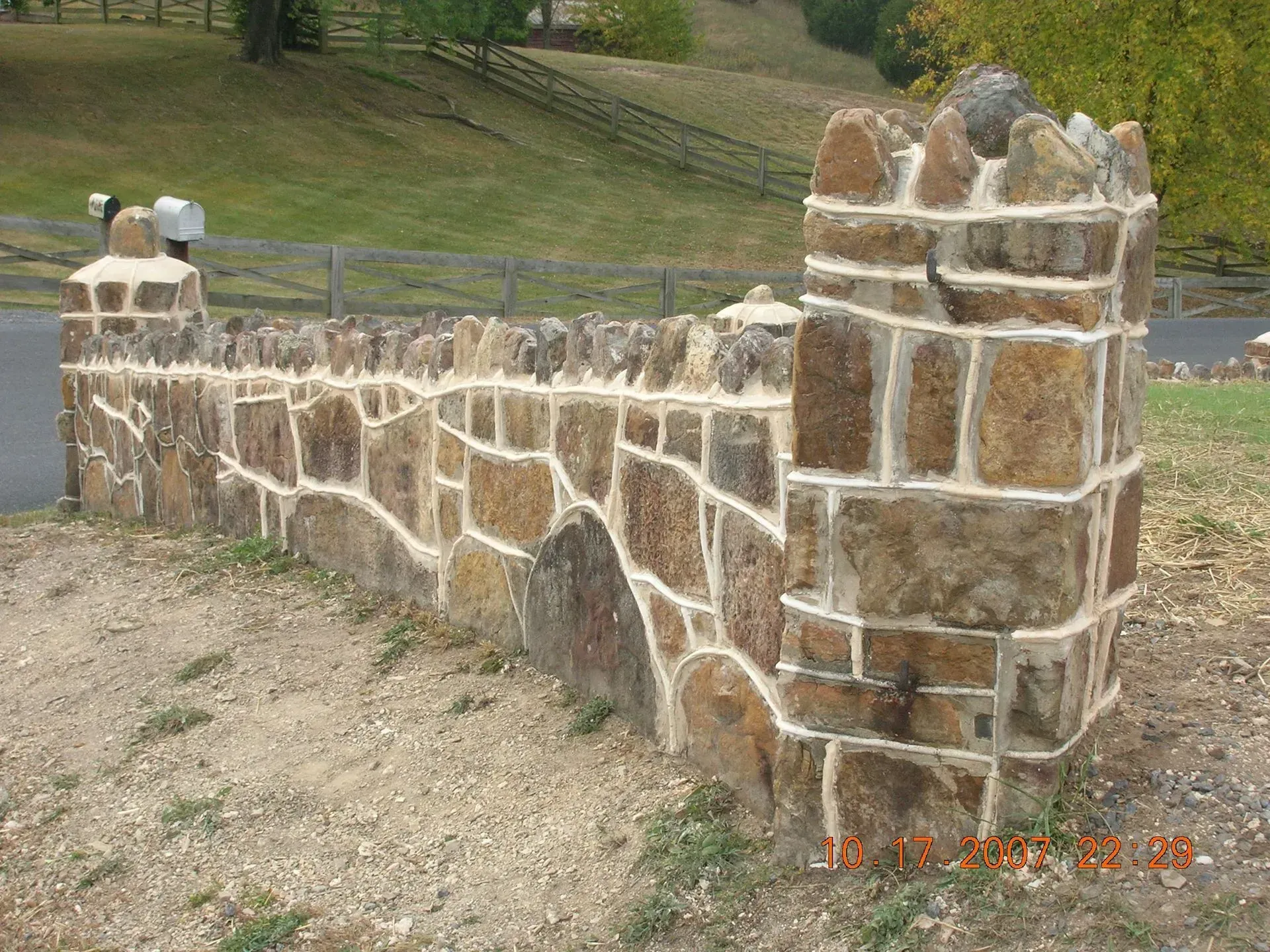 Stone wall with turreted corner and mailbox, set in a grassy area near a fence and road.