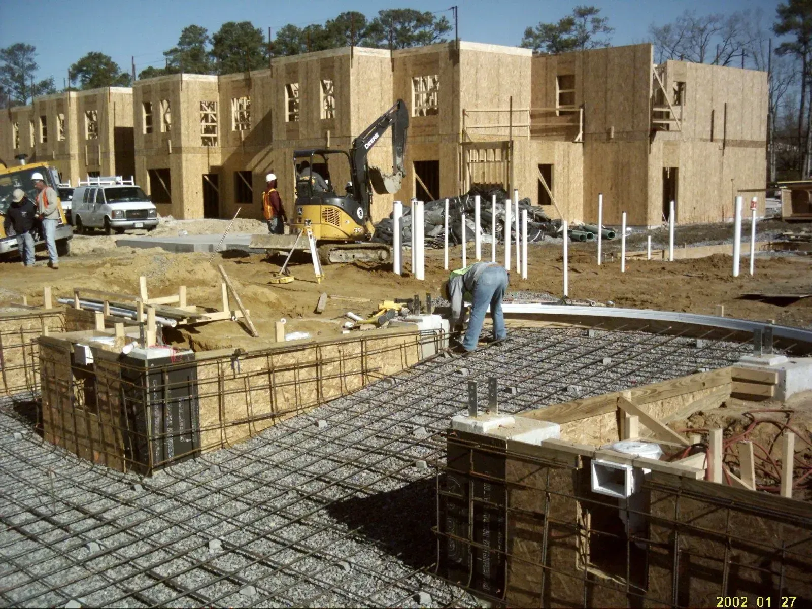 Construction site with wooden framed buildings. Workers, excavator, and concrete forms visible.