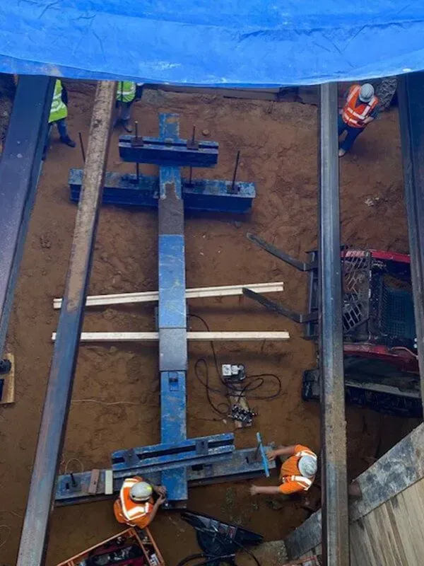 Construction workers operating machinery in a trench. Blue metal frame, orange vests, dirt, and a blue tarp overhead.