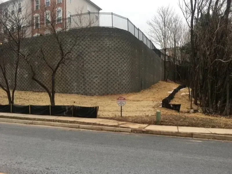 A tall, gray retaining wall with a fence sits above a grassy area next to a road.