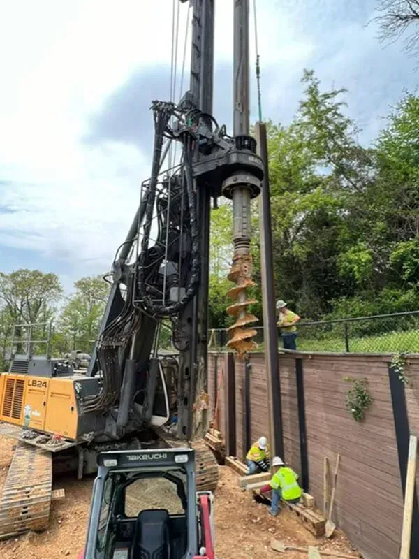 Large drilling machine excavating near a retaining wall, workers in helmets nearby, cloudy day.