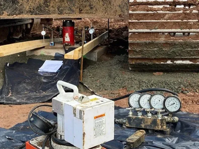 Hydraulic load test on a construction site, with gauges, a pump, and a jack under an excavator.