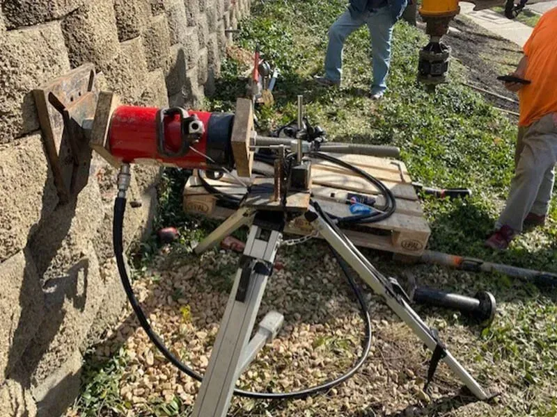 A hydraulic drill on a tripod against a retaining wall; workers nearby.