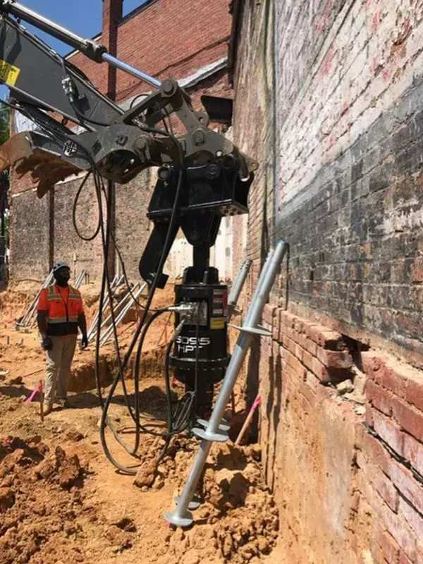 An excavator drills a support pier next to a brick building with a construction worker nearby.