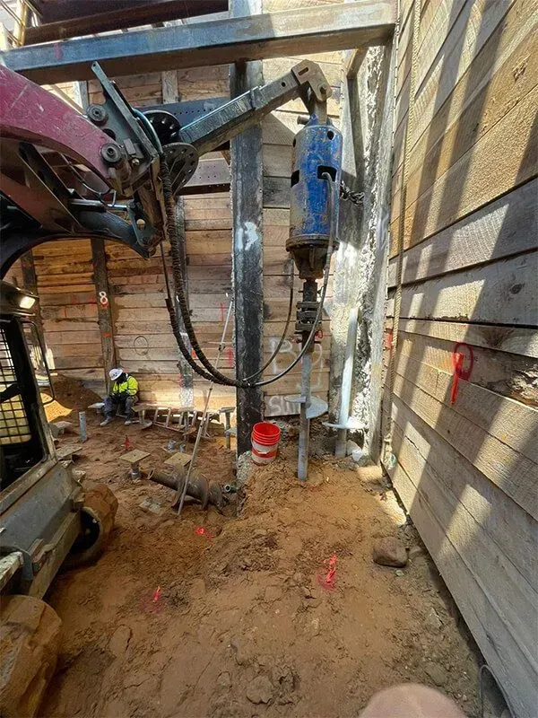 Construction site: drill rig against wooden wall, worker, red flags, and dirt.