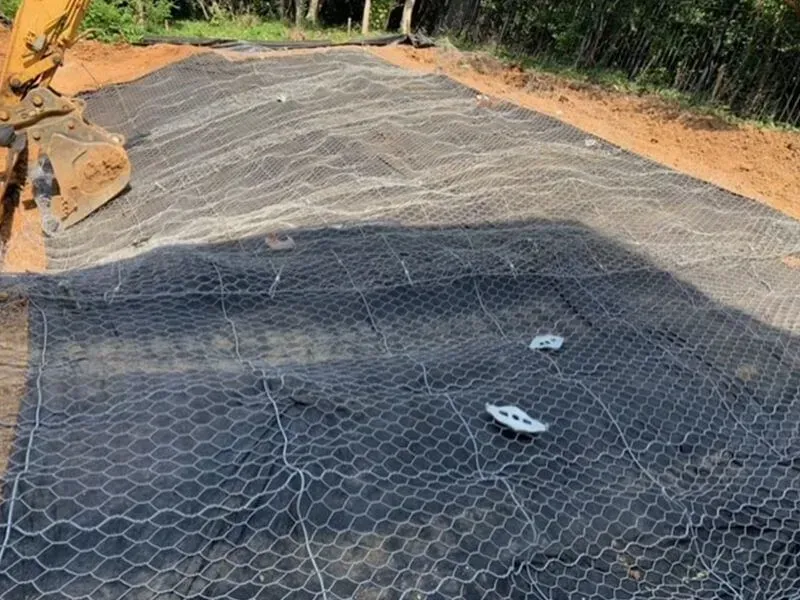 Gray wire mesh covers a black fabric on a hillside. Excavator visible in the upper left.