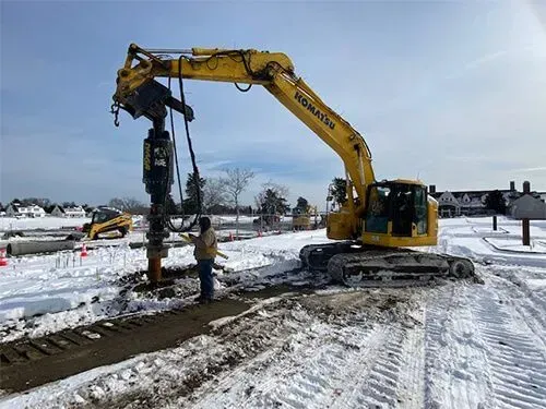 Yellow Komatsu excavator drilling in snowy construction site; worker nearby.