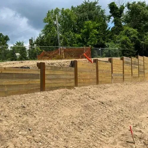 A wooden retaining wall on a dirt slope. Trees and a fence are in the background under a cloudy sky.