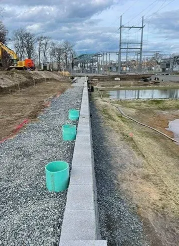 Construction site with a concrete wall, green pipes, and gravel next to a body of water.