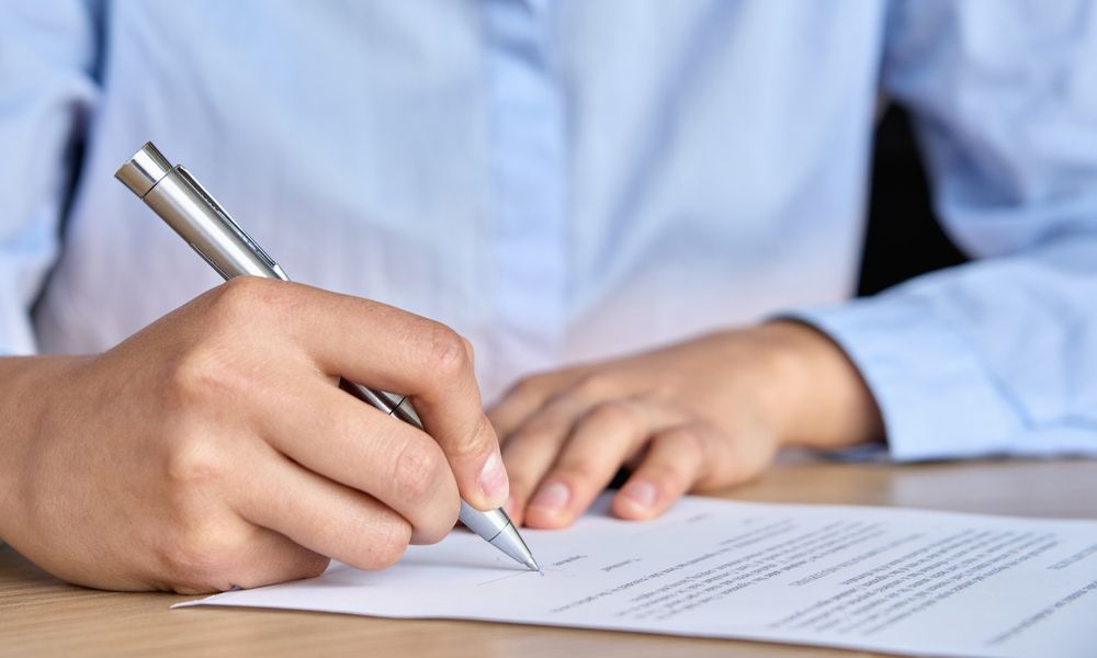 Person in blue shirt signing a document with a silver pen on a wooden desk.