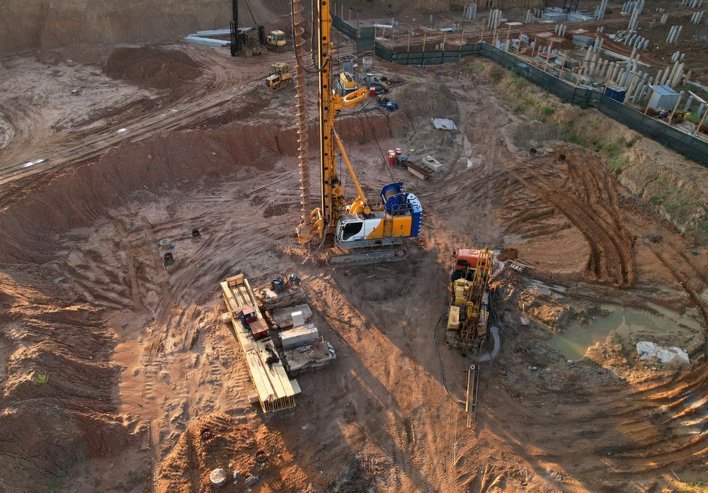 Construction site with drilling equipment. Excavated area, machinery, and foundation framework visible.
