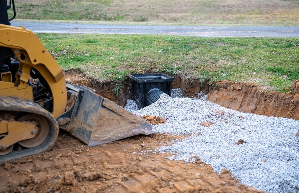 Yellow excavator at a ditch with gravel, next to a black drainage box in a dirt opening near a road and grass.