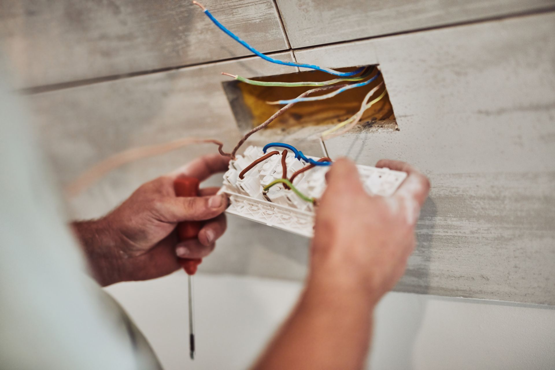 Hands connecting electrical wires in wall box opening on tiled wall.
