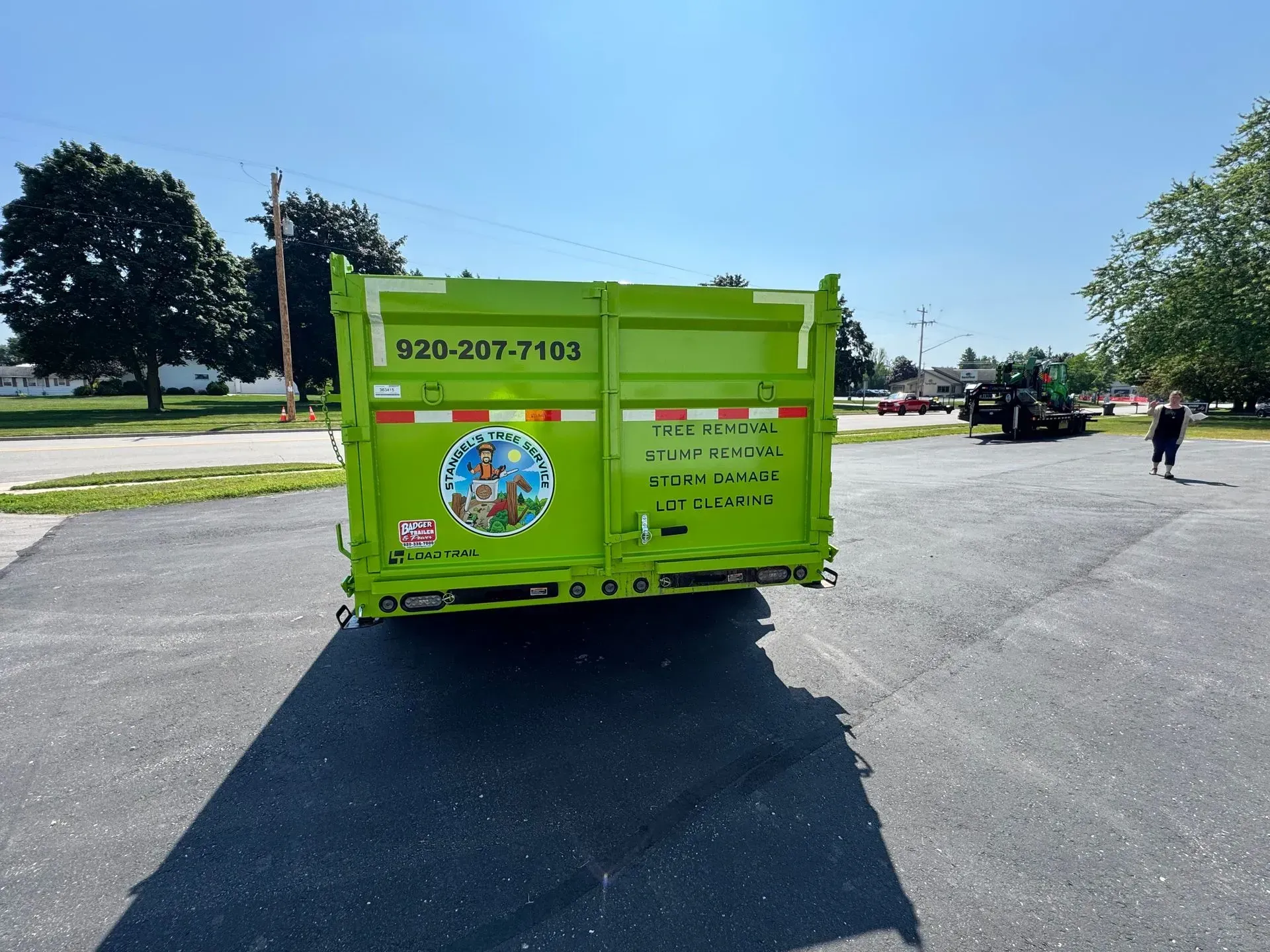 Bright green dumpster truck parked on a sunny street with trees and a person in the distance