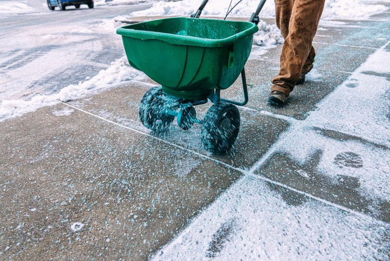 Green salt spreader on a snowy sidewalk with a person walking beside it