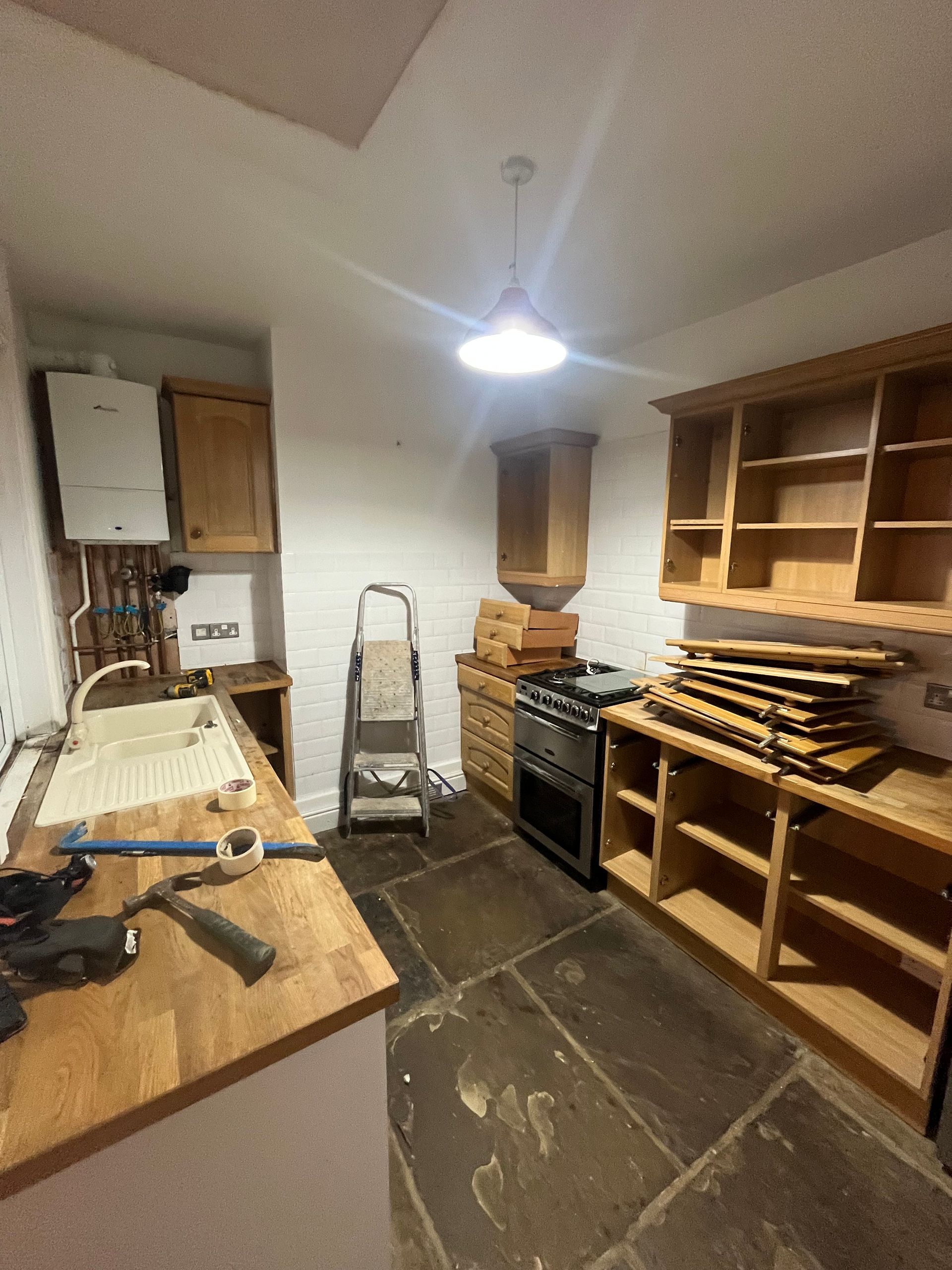 Kitchen renovation: cabinets, oven, sink, and appliances, in progress. Stone floor, white walls, and ladder visible.