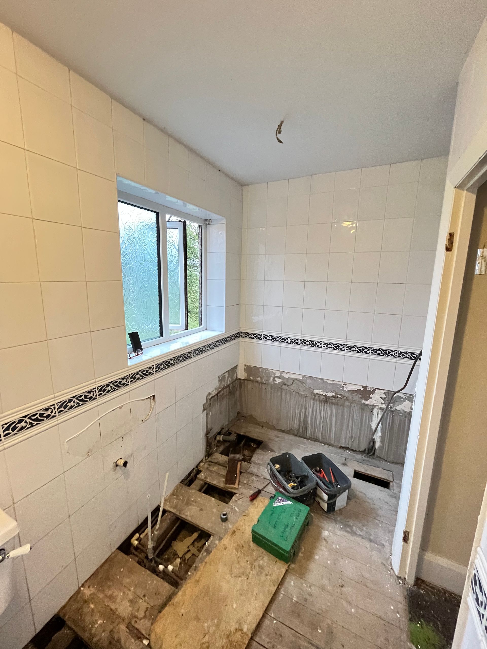 Bathroom under renovation, with exposed floorboards, tiled walls, and a window. Tools visible on the floor.