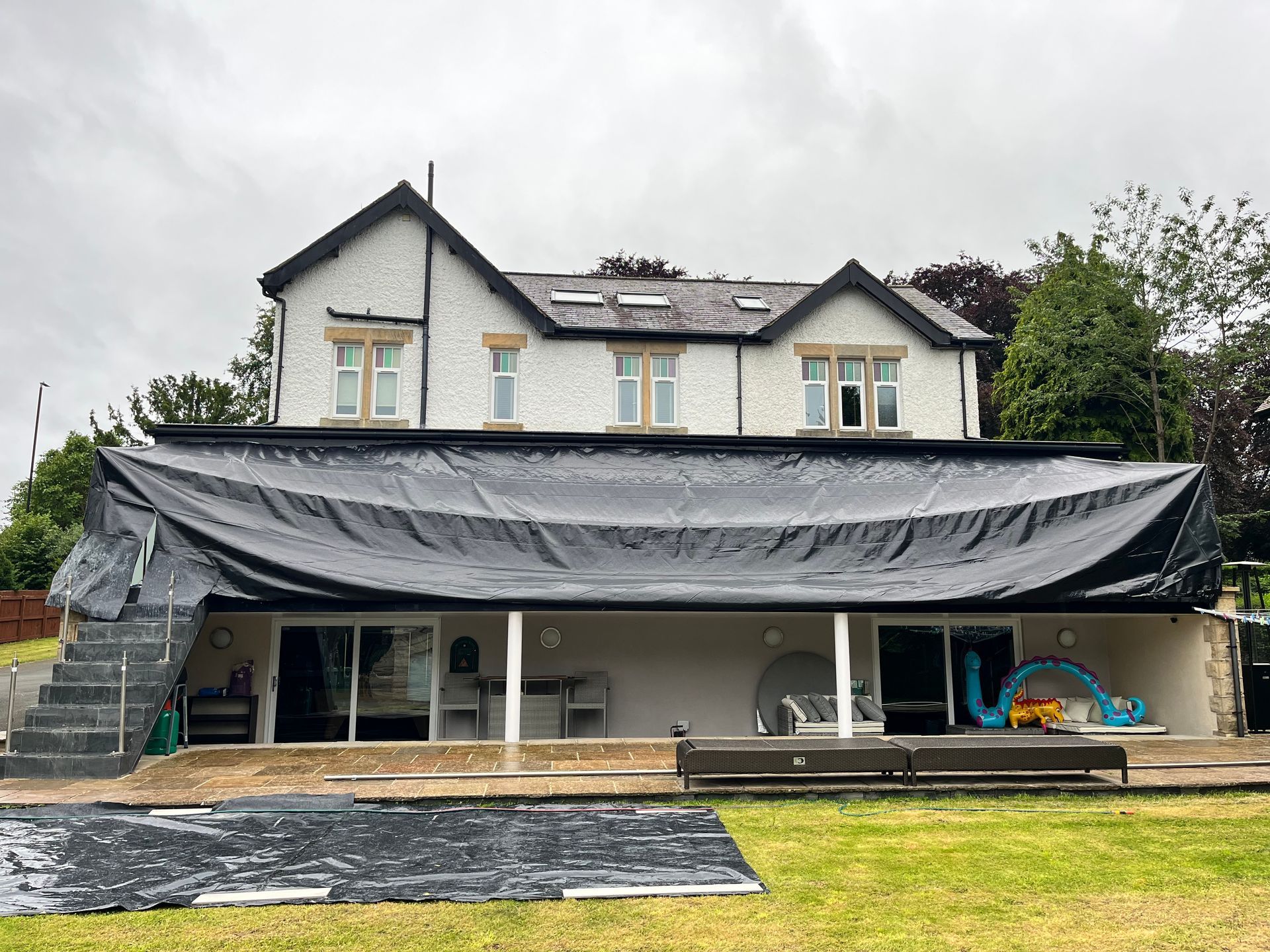 White house with a black tarp covering a lower level, gray skies, outdoor setting.