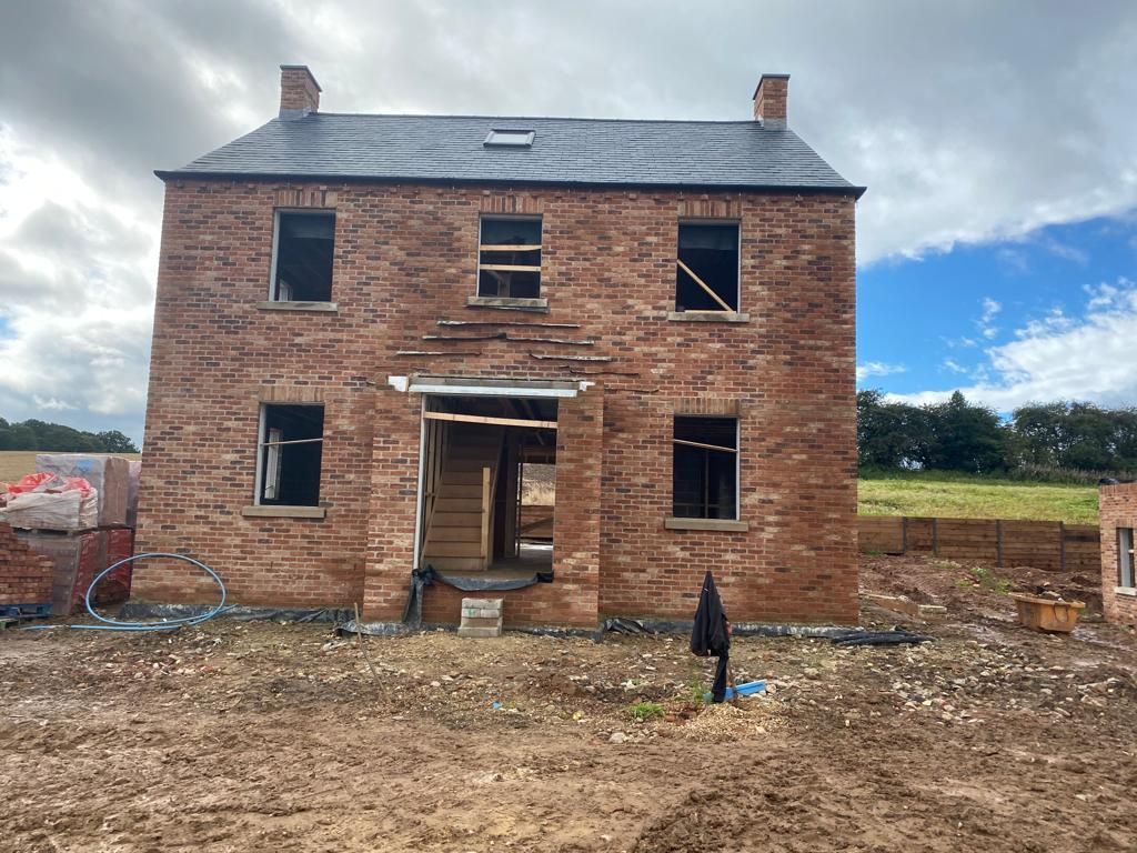 Brick house under construction, with open windows, a slated roof, and surrounding muddy ground.