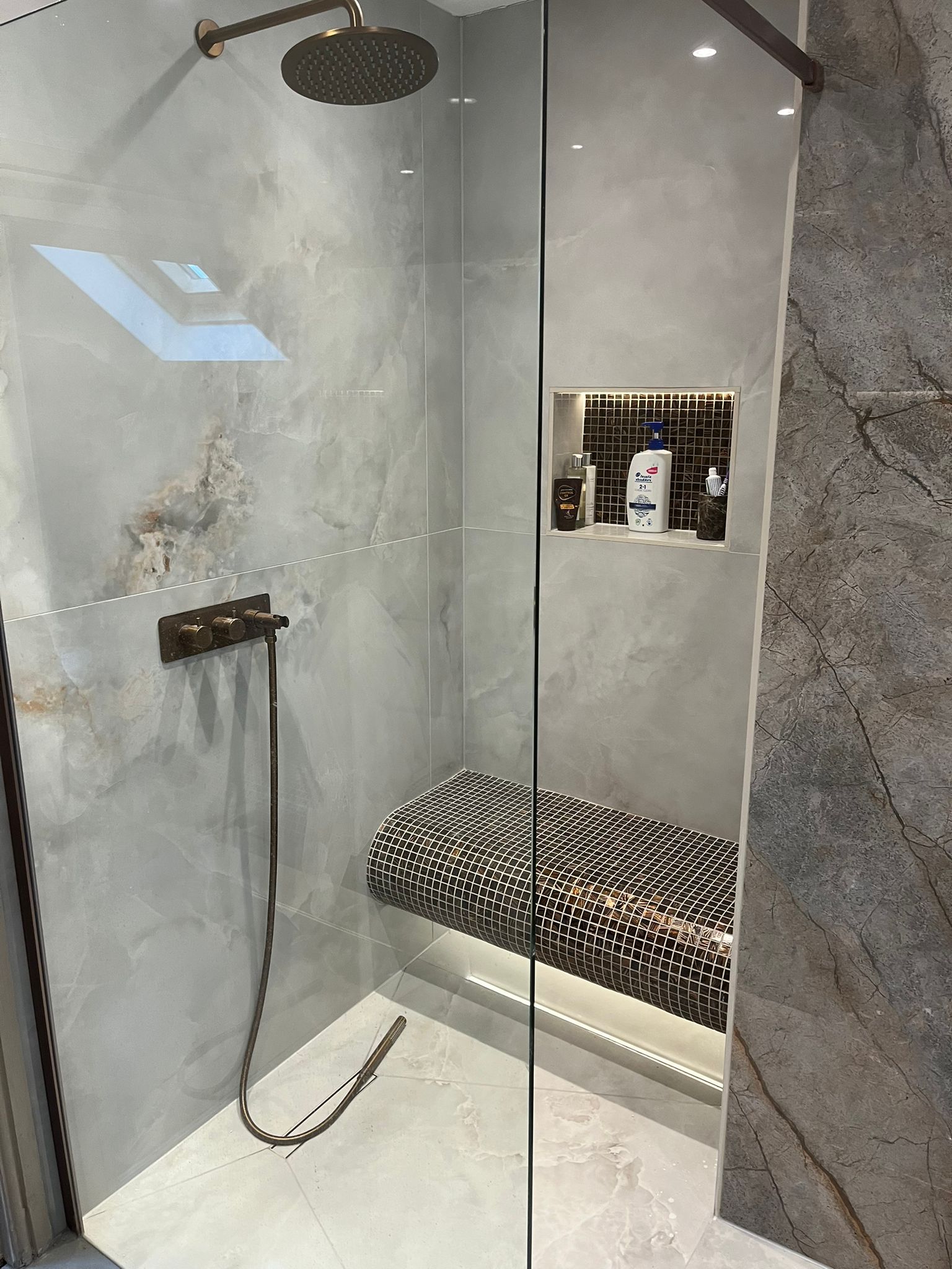 Shower with grey marble-look tiles, built-in bench, recessed shelf with products, and bronze fixtures.