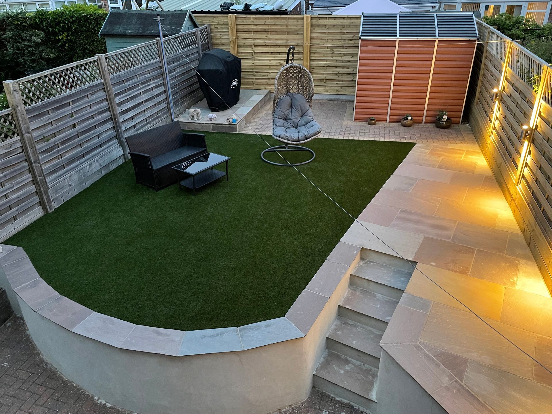 Backyard patio with artificial grass, seating area, shed, and stone steps illuminated by string lights.