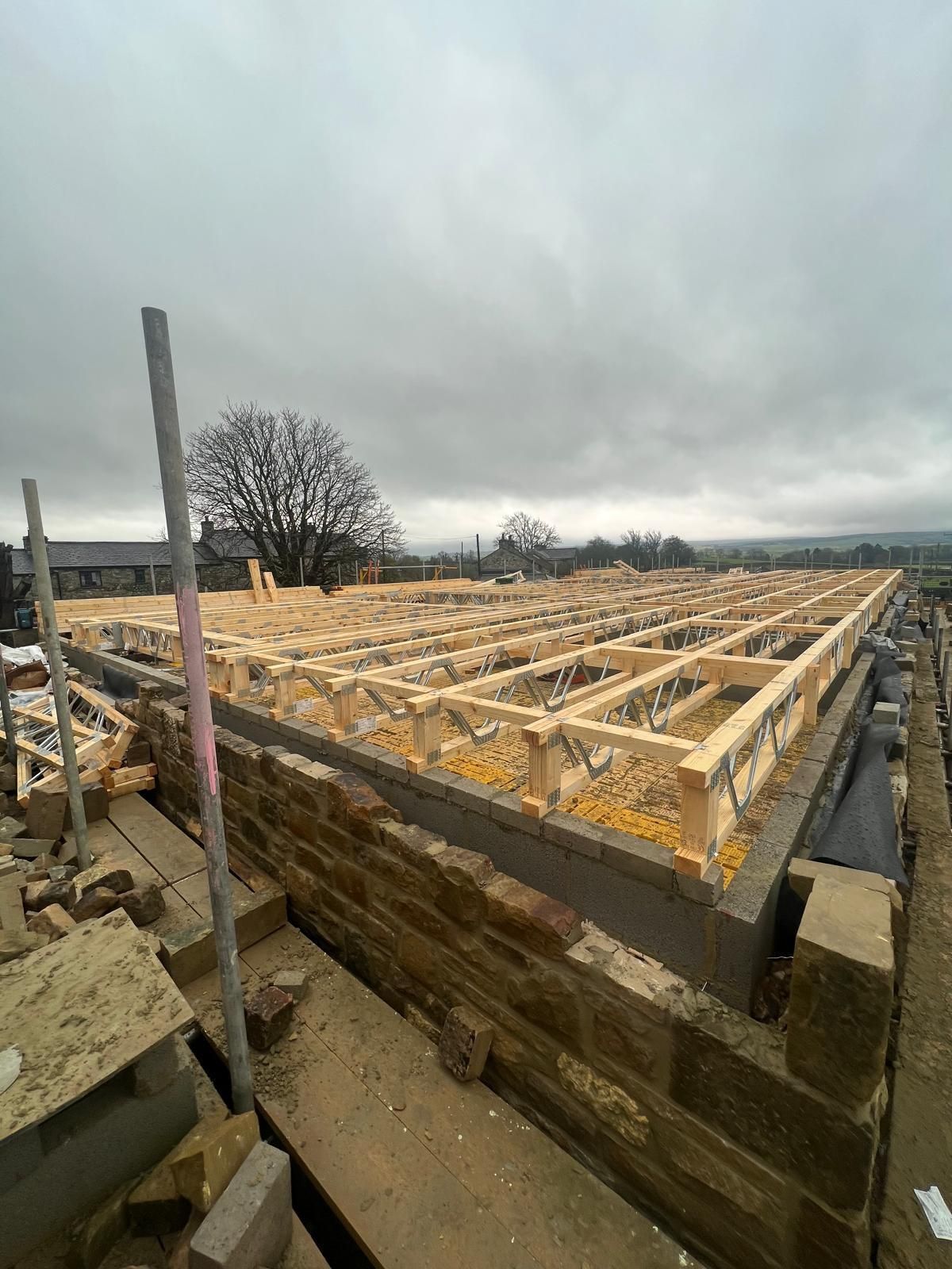 Construction site, partially built wooden structure with stone foundation, overcast sky.