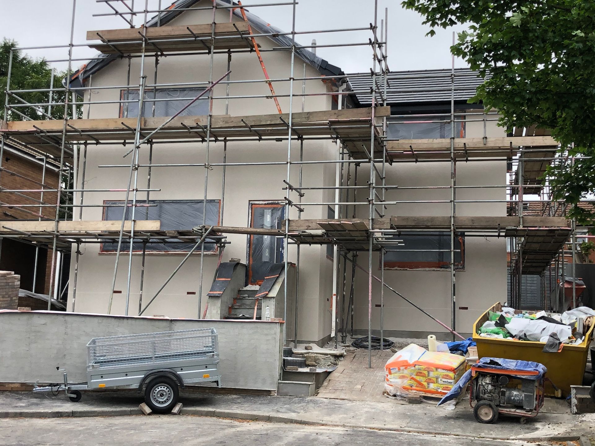 House exterior under renovation with scaffolding. Cream-colored walls, gray roof. Materials and trailer present.