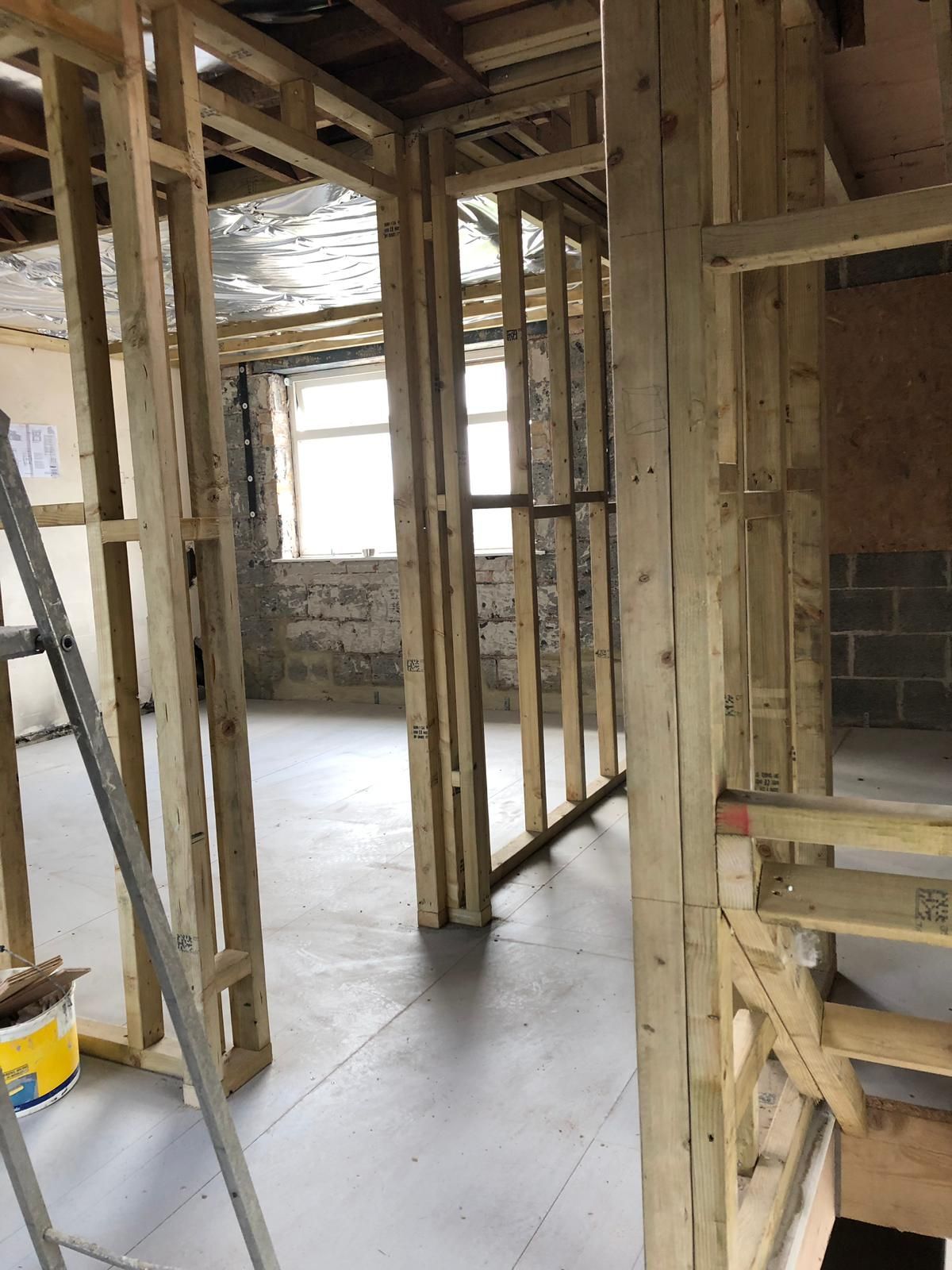 Interior framing of a building under construction with wooden studs, ladder, and gray floor.
