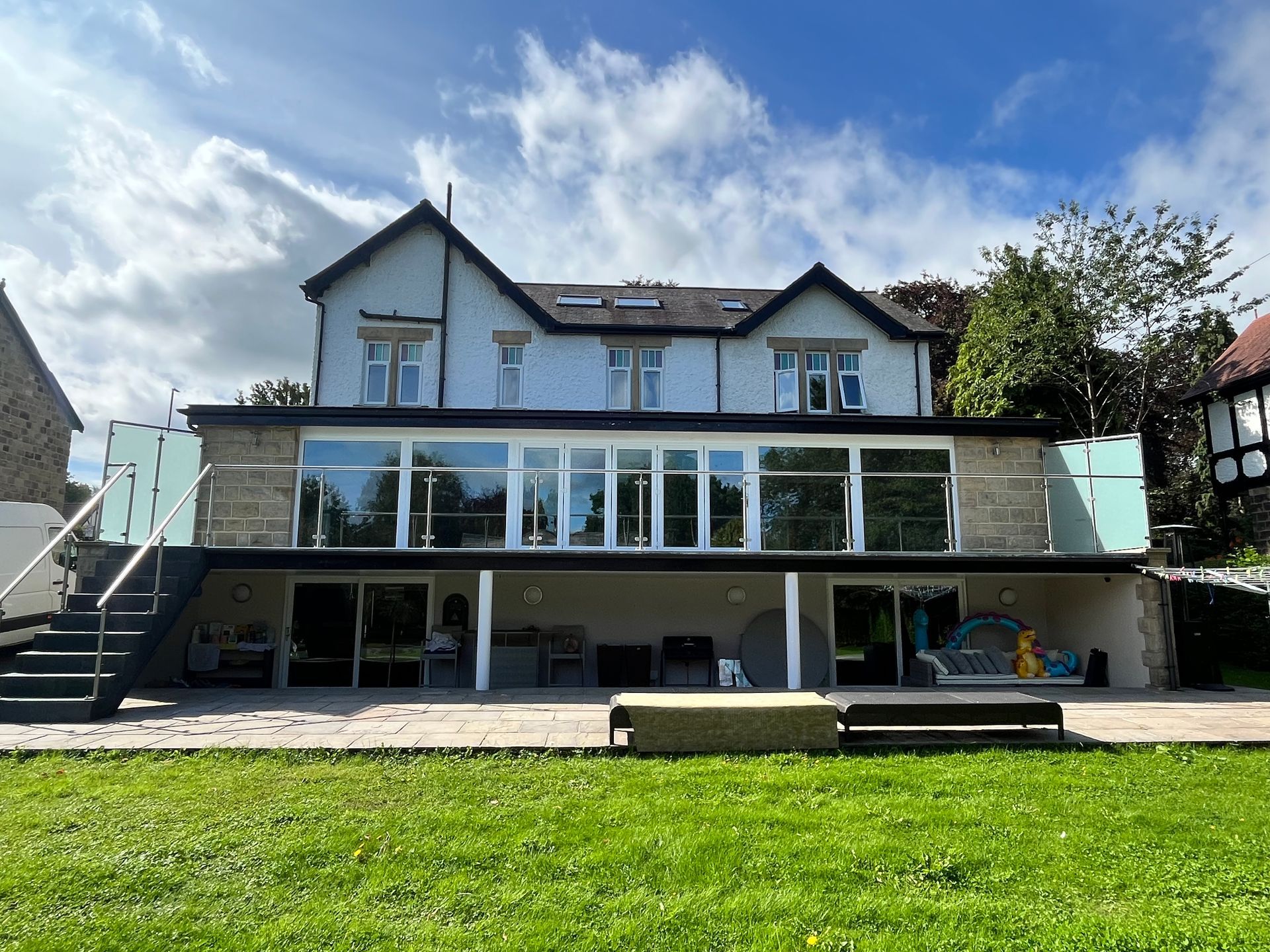Back view of a light blue house with a glass balcony and patio, set on a green lawn under a blue sky.