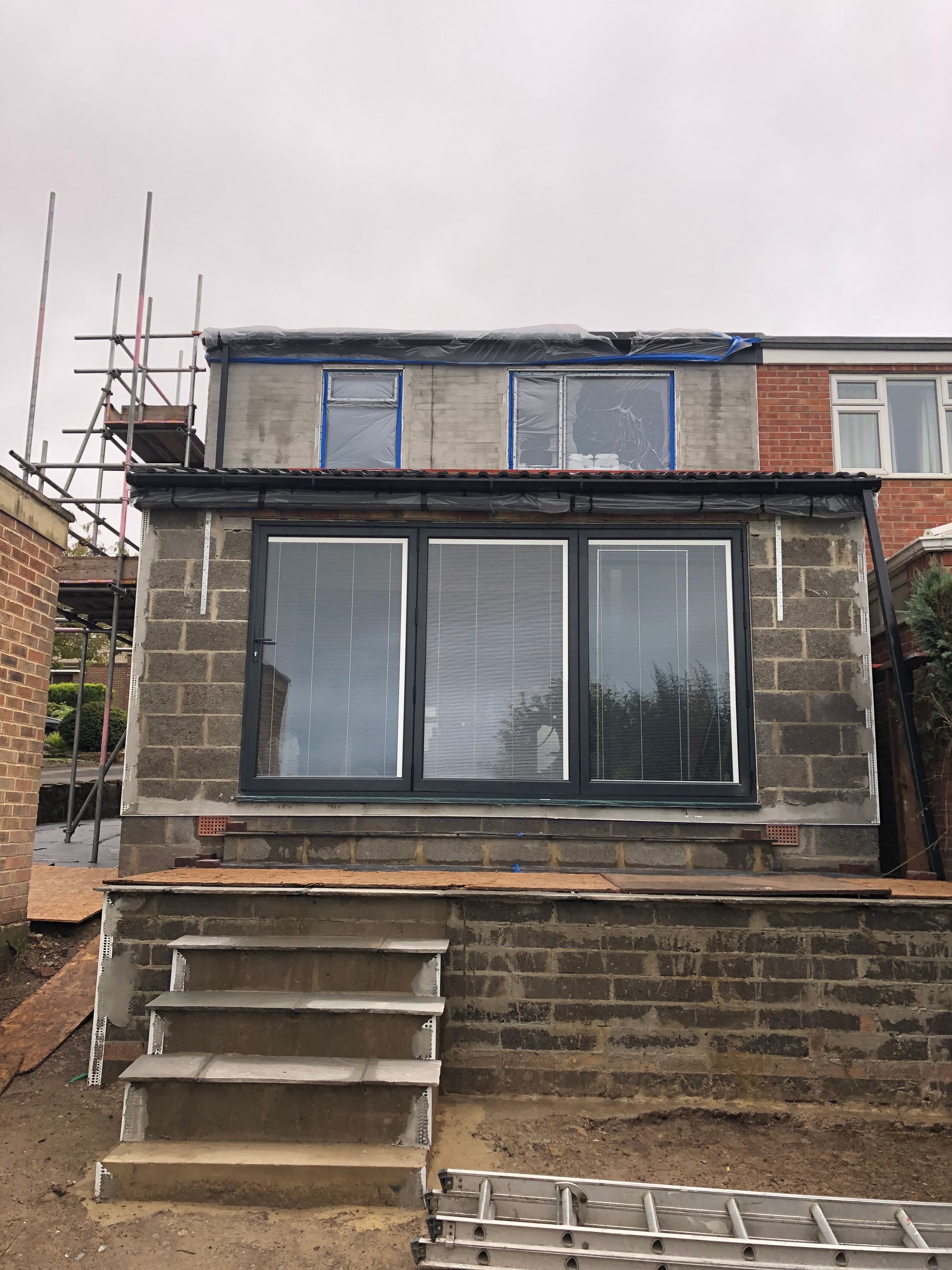 Construction in progress: extension with gray cinder block walls, windows, scaffolding, and steps. Overcast sky.