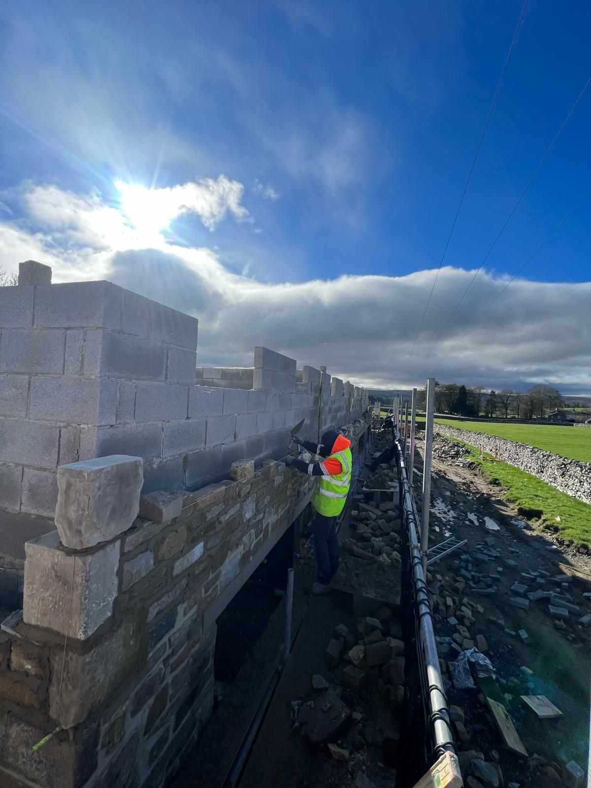 Construction worker building a stone wall on a sunny day. Blocks and stones are visible, with blue sky and clouds.