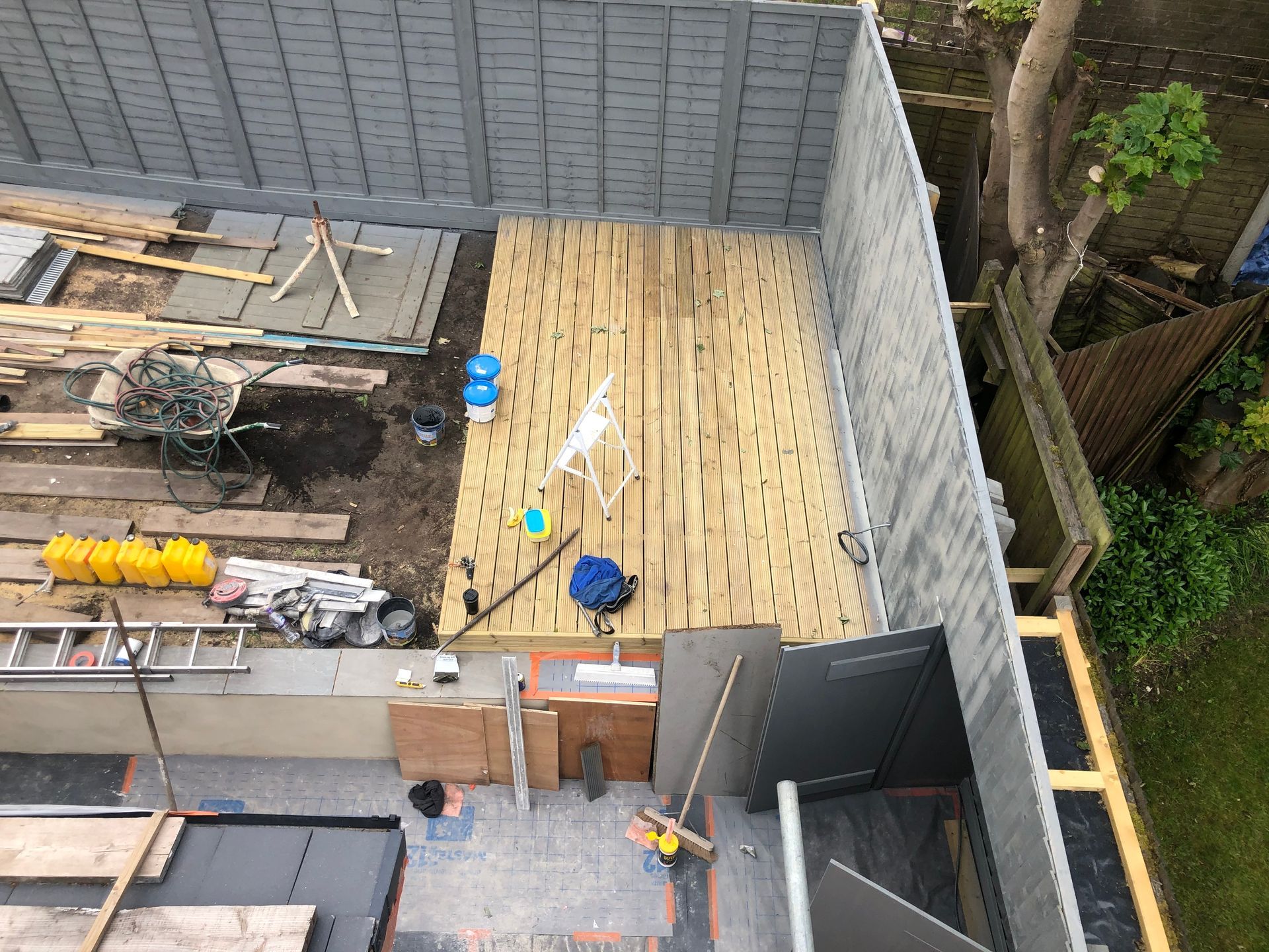 Overhead shot of a backyard with a partially built wooden deck and construction materials; gray fence.