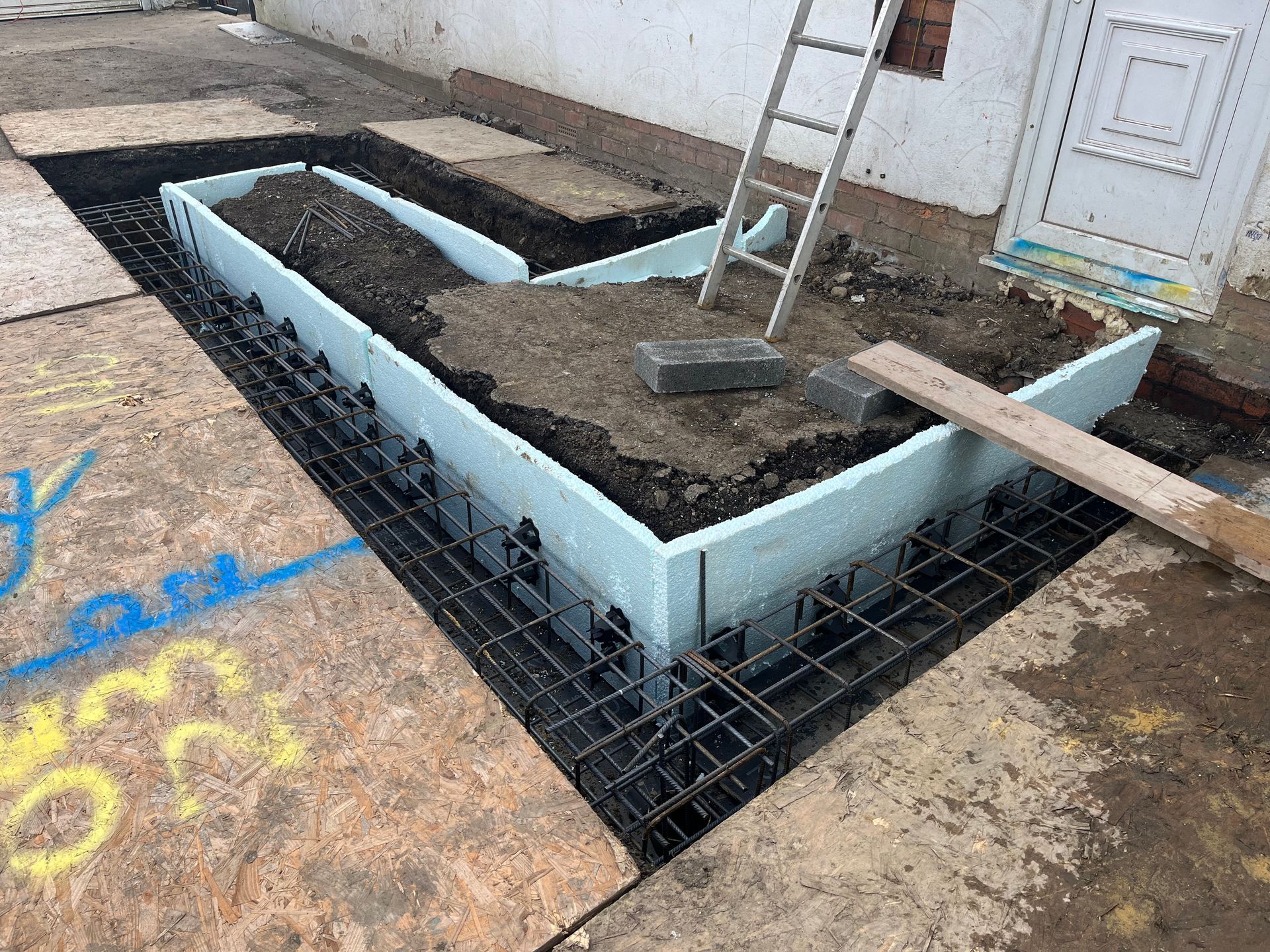 Construction site: Foam forms and rebar for foundation of building. Brown ground, white walls, ladder.