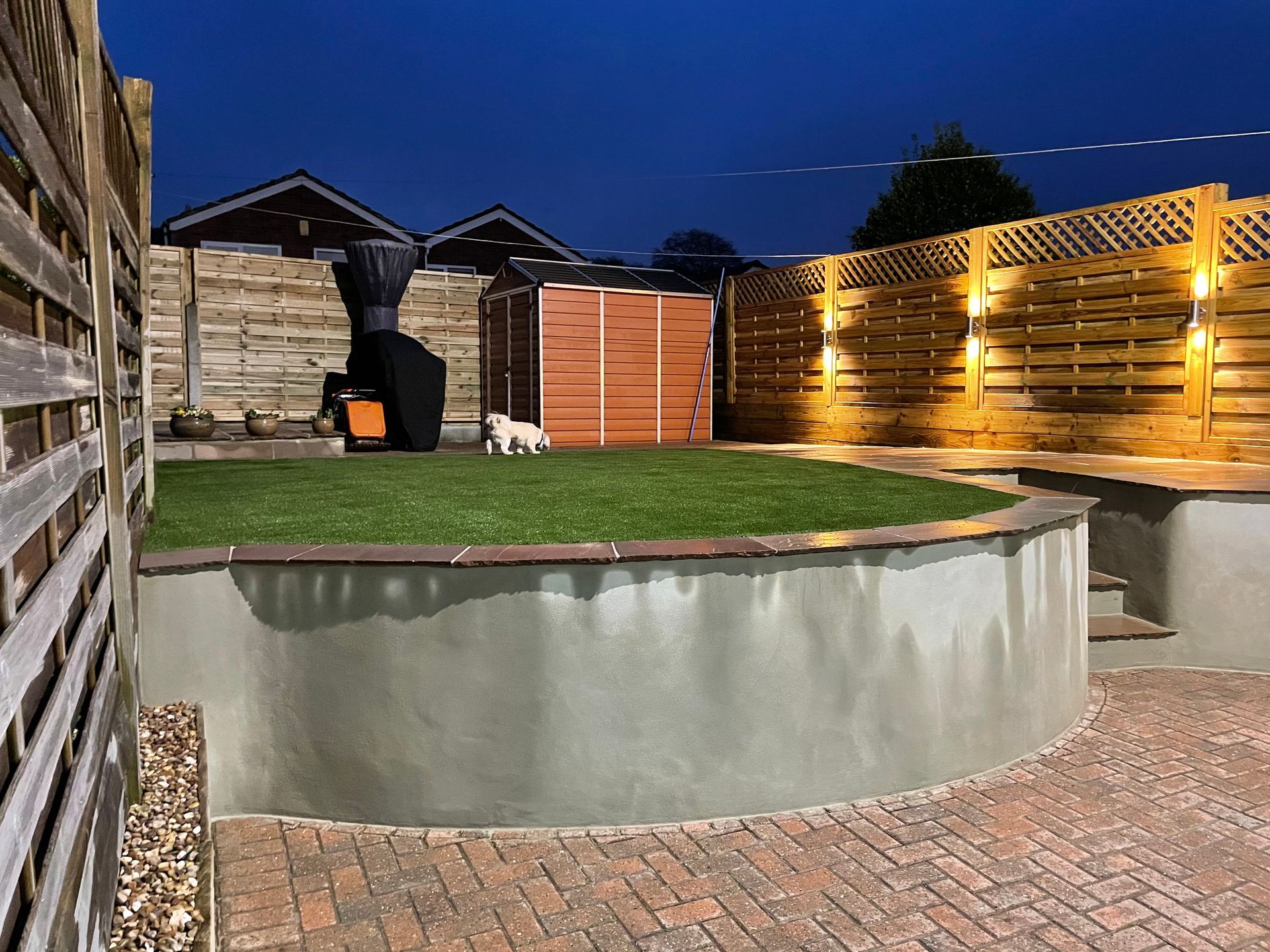 Nighttime backyard with brick patio, curved retaining wall, green turf, wooden fence, and string lights.