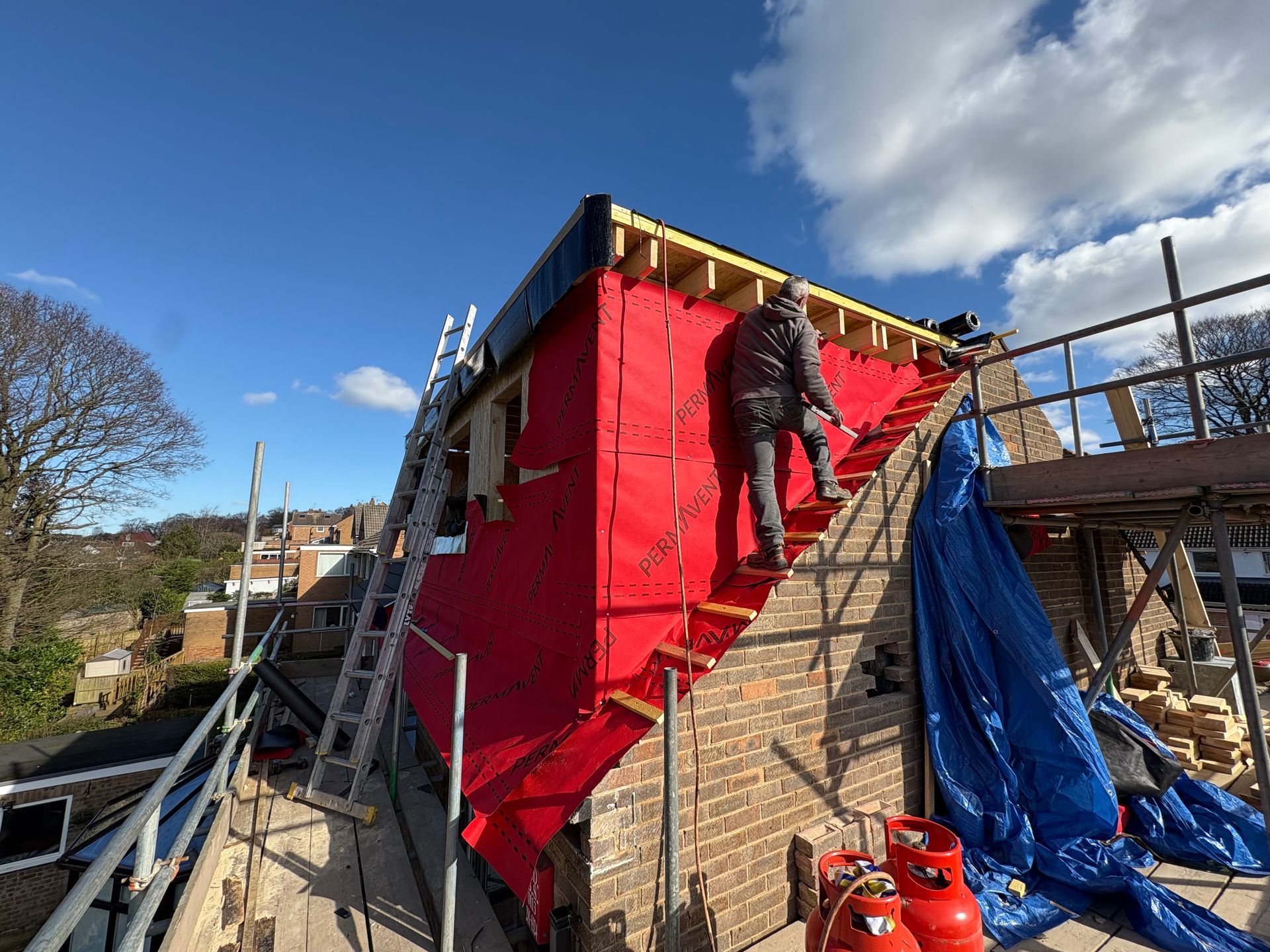 A worker on a roof covered in red material, using a ladder, under a blue sky.