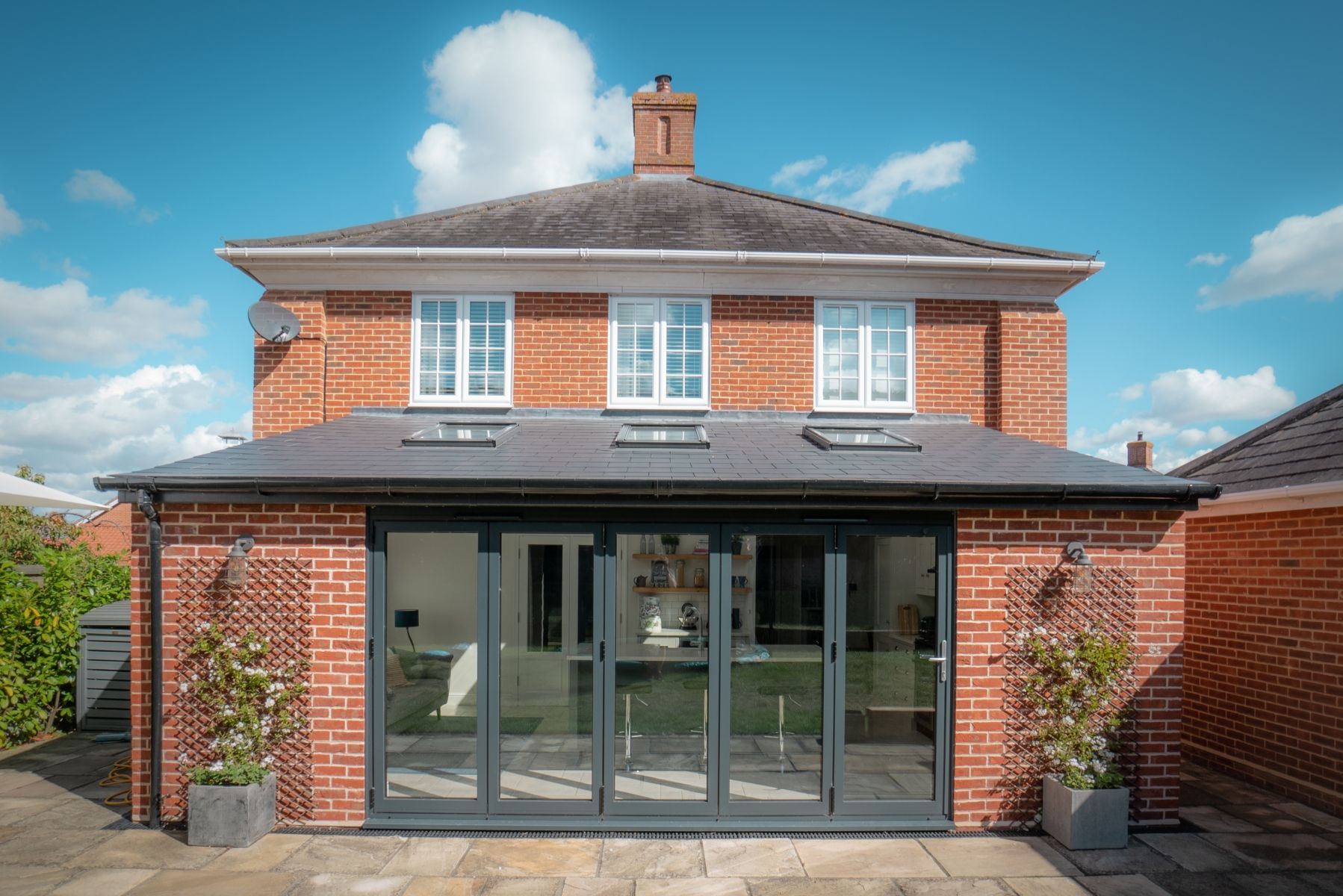 Red brick house with a rear extension featuring gray folding doors and two potted plants.