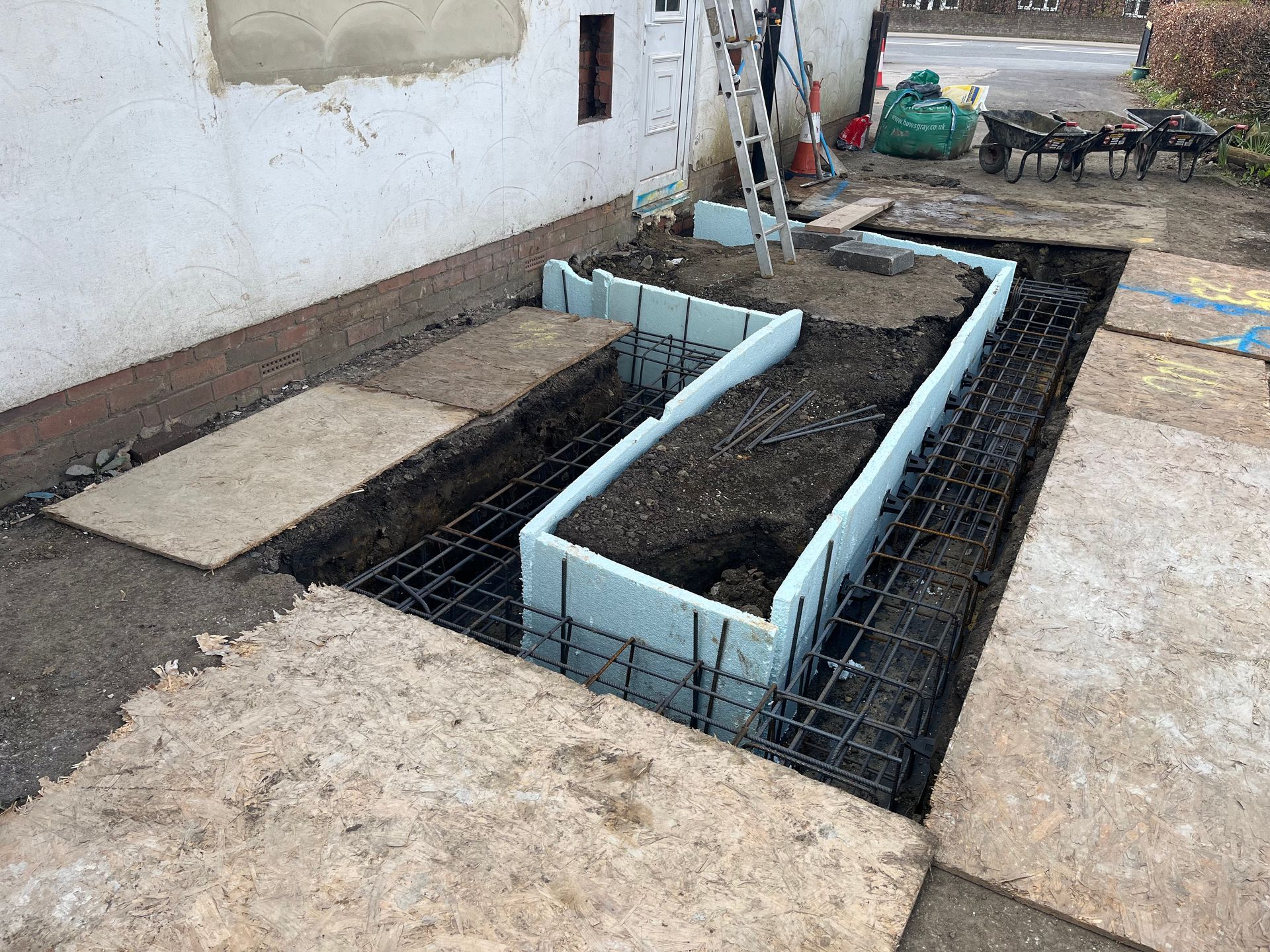 Construction site: foam forms for foundation walls, rebar grid in trench near a building.
