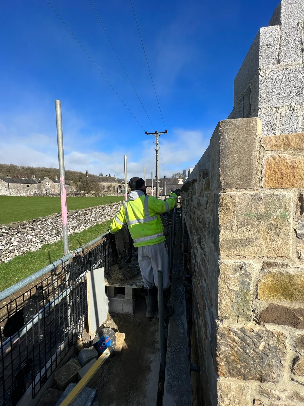 Person in safety gear walking on narrow ledge of stone wall. Rural setting with a blue sky.