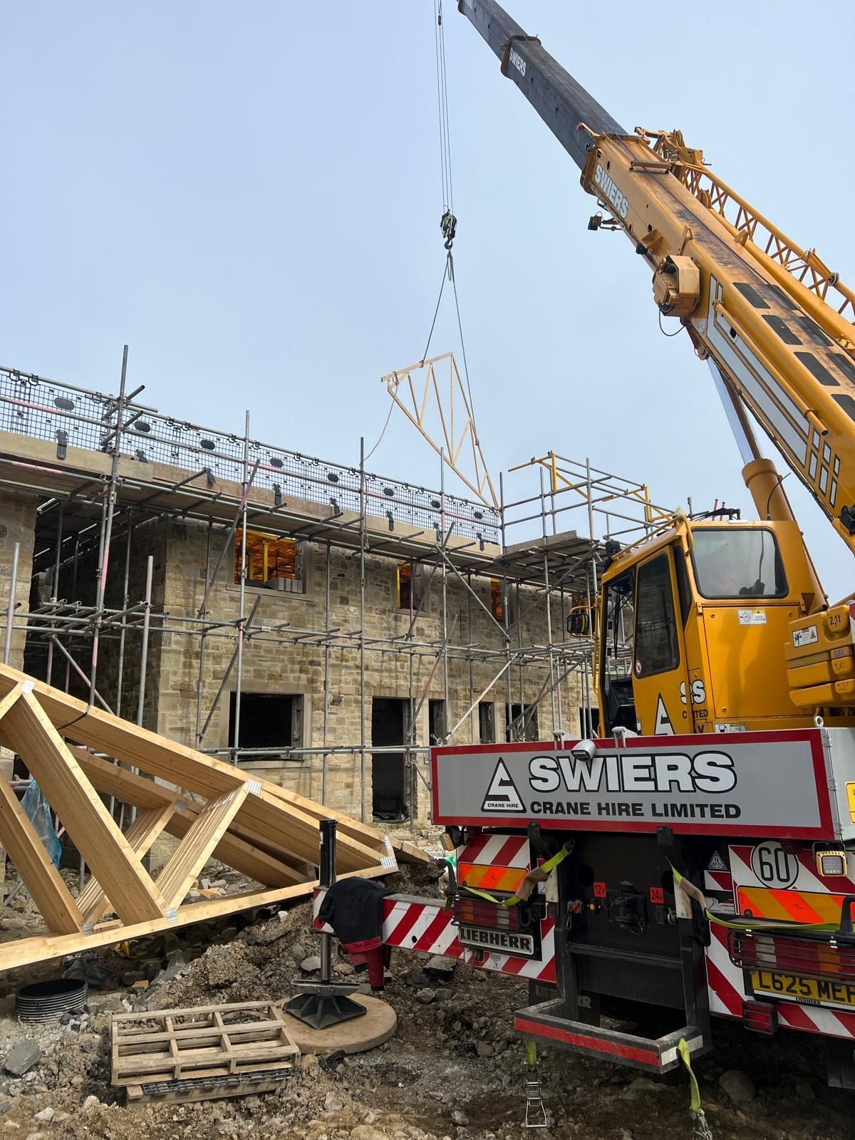 A crane lifting a wooden roof truss onto a building under construction. Workers are present.