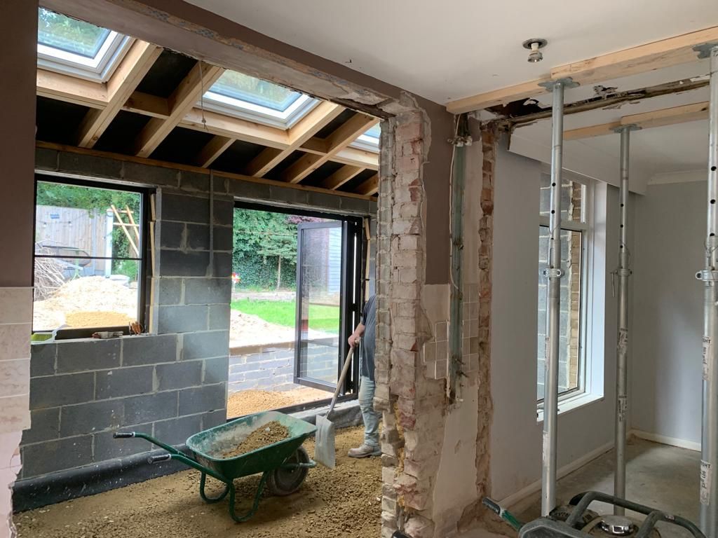 Interior renovation: wall demolition in progress, revealing brick and steel supports; wheelbarrow holds debris.