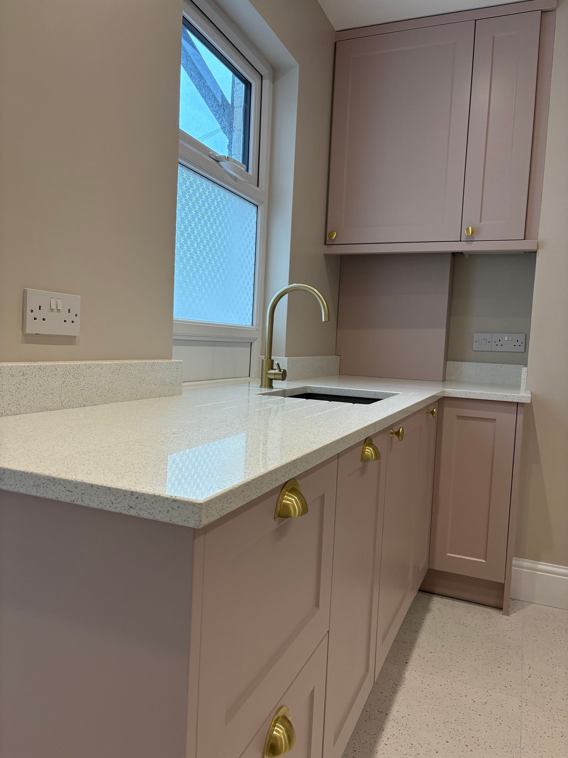 Pink kitchen cabinets with gold hardware, quartz countertop, and a window.