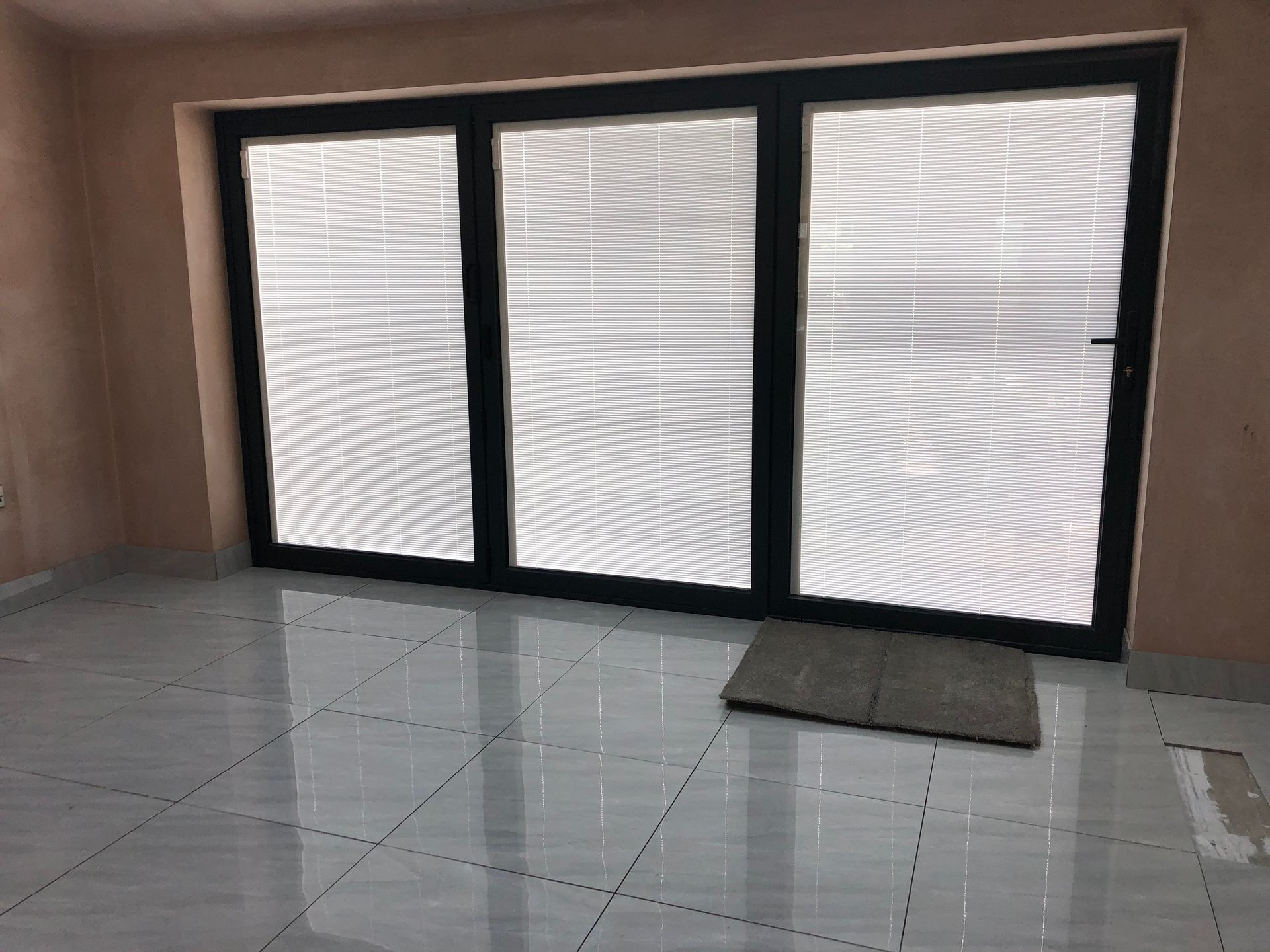Large black framed window with three panels, with white blinds, in a room with tiled floor and pink walls.